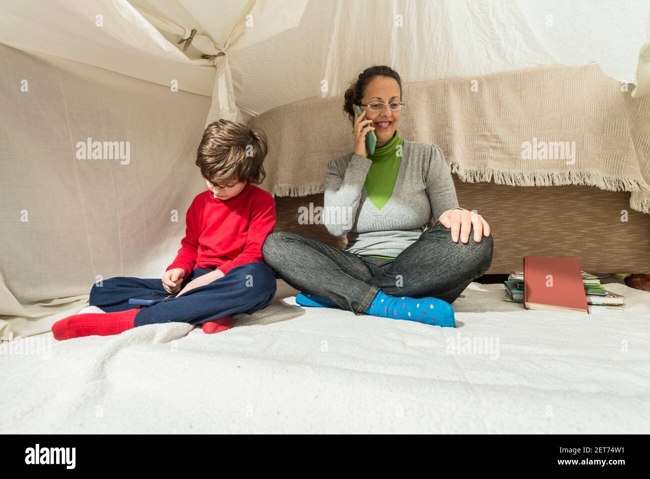 A woman talks on the phone and a child plays with a smartphone in a makeshift tent in their living room. Home lifestyle concept. Stock Photo