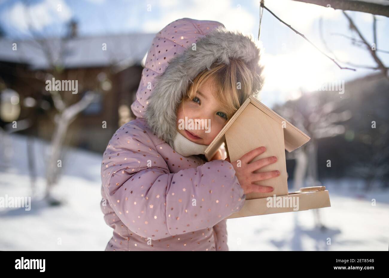 Small girl outdoors in winter garden, standing by wooden bird feeder. Stock Photo