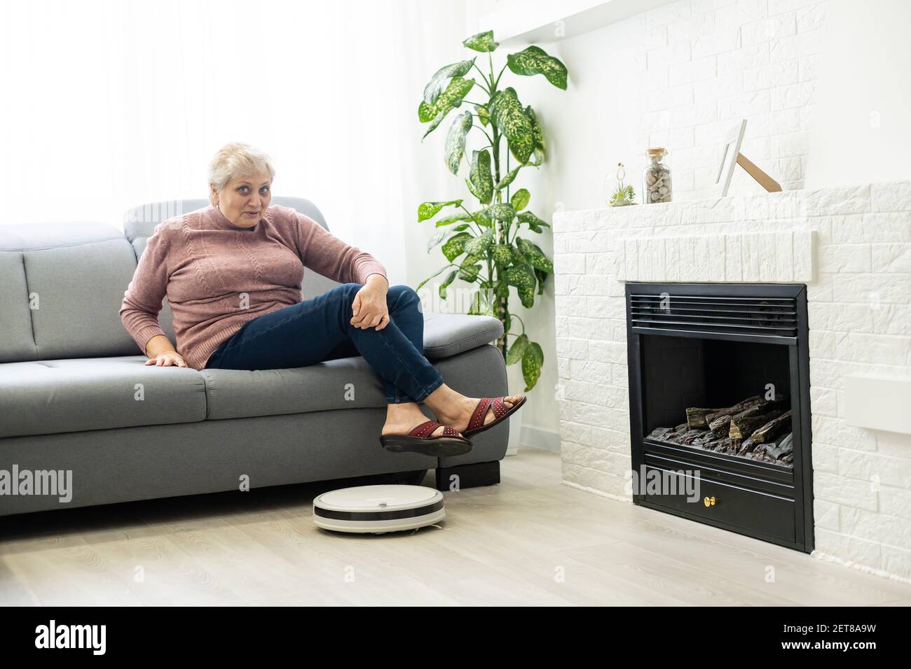 An elderly woman uses a robot vacuum cleaner in an apartment Stock
