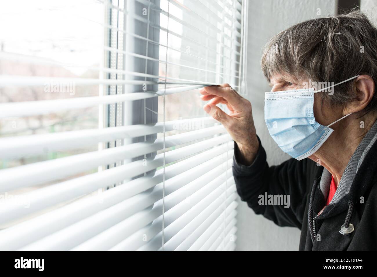 An elderly woman in a medical mask looks out the window. Stock Photo