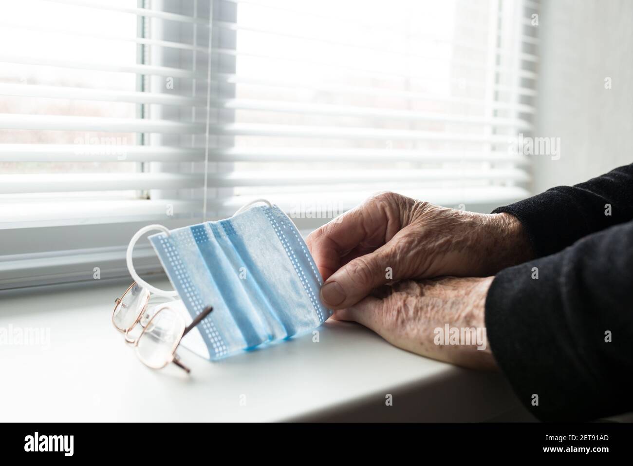 Old human hands holding medical mask next to the window, on self-isolation. Stock Photo