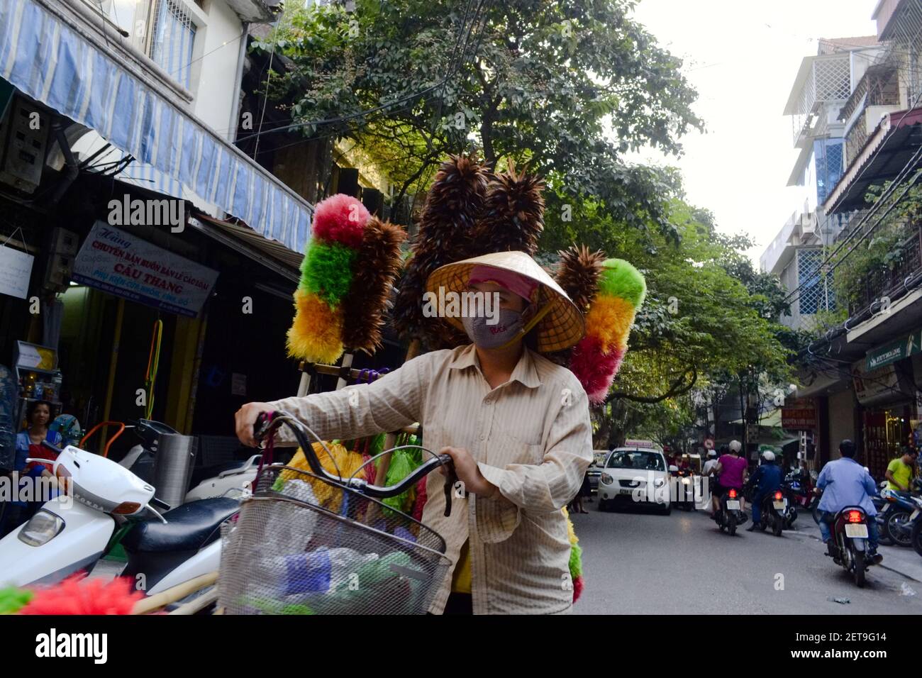 Hanoi, Vietnam - September, 2015: Vietnamese woman in mask walking with bike and selling colorful feather dusters on the city street Stock Photo