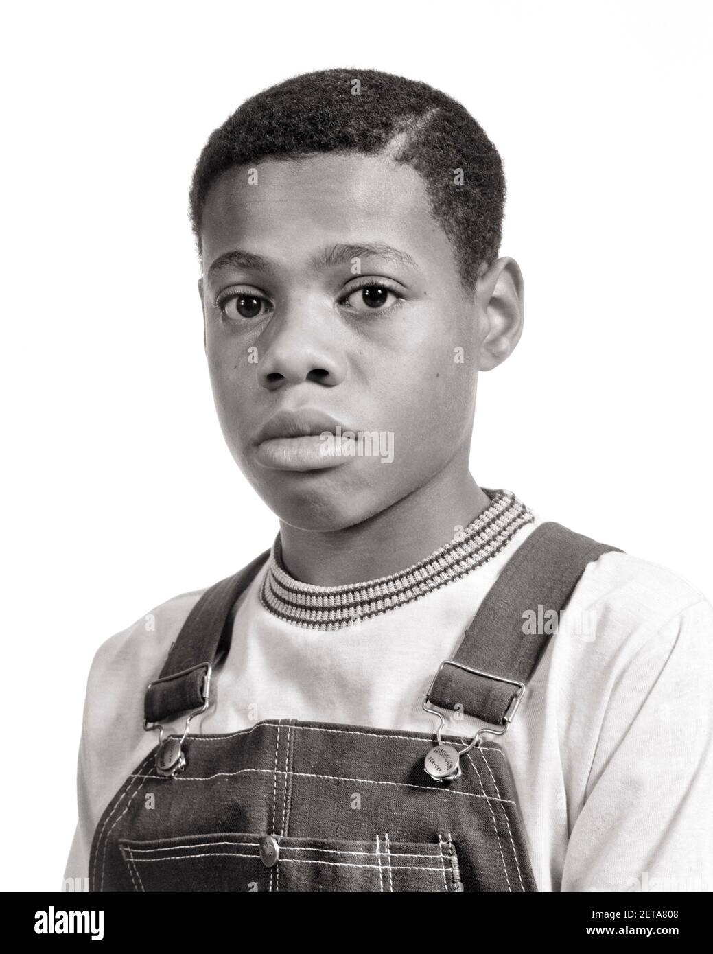 1970s PORTRAIT OF PRE-TEEN AFRICAN-AMERICAN BOY WEARING DENIM BIB OVERALLS SERIOUS FACIAL EXPRESSION LOOKING AT CAMERA  - b25648 HAR001 HARS SADNESS EYE CONTACT HEAD AND SHOULDERS AFRICAN-AMERICANS AFRICAN-AMERICAN BLACK ETHNICITY TEENAGED SINCERE SOLEMN BIB FOCUSED INTENSE JUVENILES PRE-TEEN PRE-TEEN BOY BLACK AND WHITE CAREFUL EARNEST HAR001 INTENT OLD FASHIONED AFRICAN AMERICANS Stock Photo