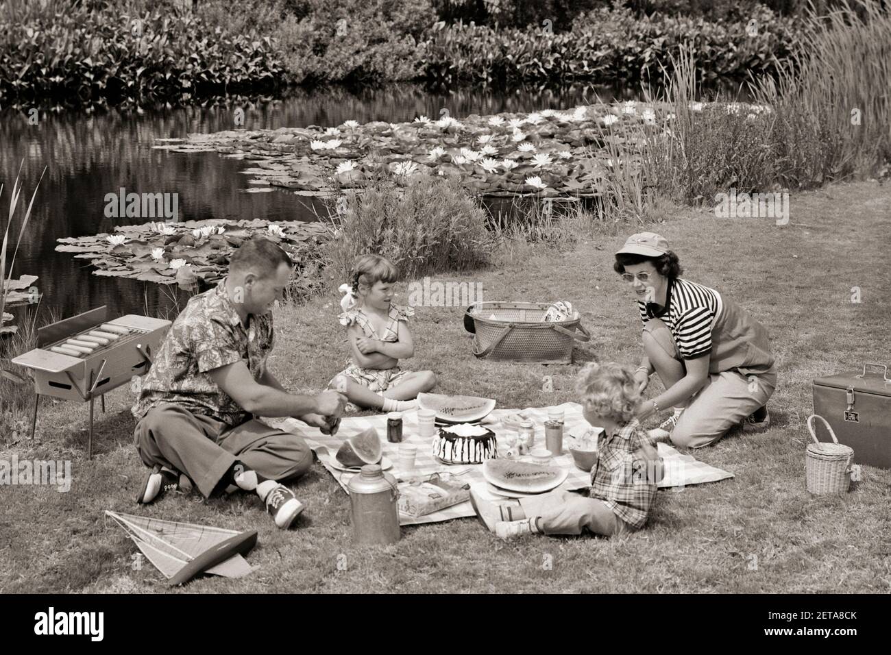 1950s FAMILY MOTHER FATHER AND TWO DAUGHTERS HAVING A SUMMER PICNIC BY POND - c174 CRS001 HARS PAIR 4 SUBURBAN SUNGLASSES MOTHERS OLD TIME NOSTALGIA OLD FASHION SISTER 1 JUVENILE STYLE POND FAMILIES JOY LIFESTYLE CELEBRATION FEMALES MARRIED RURAL SPOUSE HUSBANDS HEALTHINESS COPY SPACE FRIENDSHIP FULL-LENGTH HALF-LENGTH LADIES DAUGHTERS PERSONS SCENIC MALES SIBLINGS SISTERS FATHERS B&W PARTNER SUMMERTIME HAPPINESS HIGH ANGLE ADVENTURE LEISURE AND DADS RECREATION SIBLING WATERMELON AL FRESCO SADDLE OXFORDS PICNICKING STYLISH COOPERATION HOT DOGS JUVENILES MID-ADULT MID-ADULT MAN MID-ADULT WOMAN Stock Photo