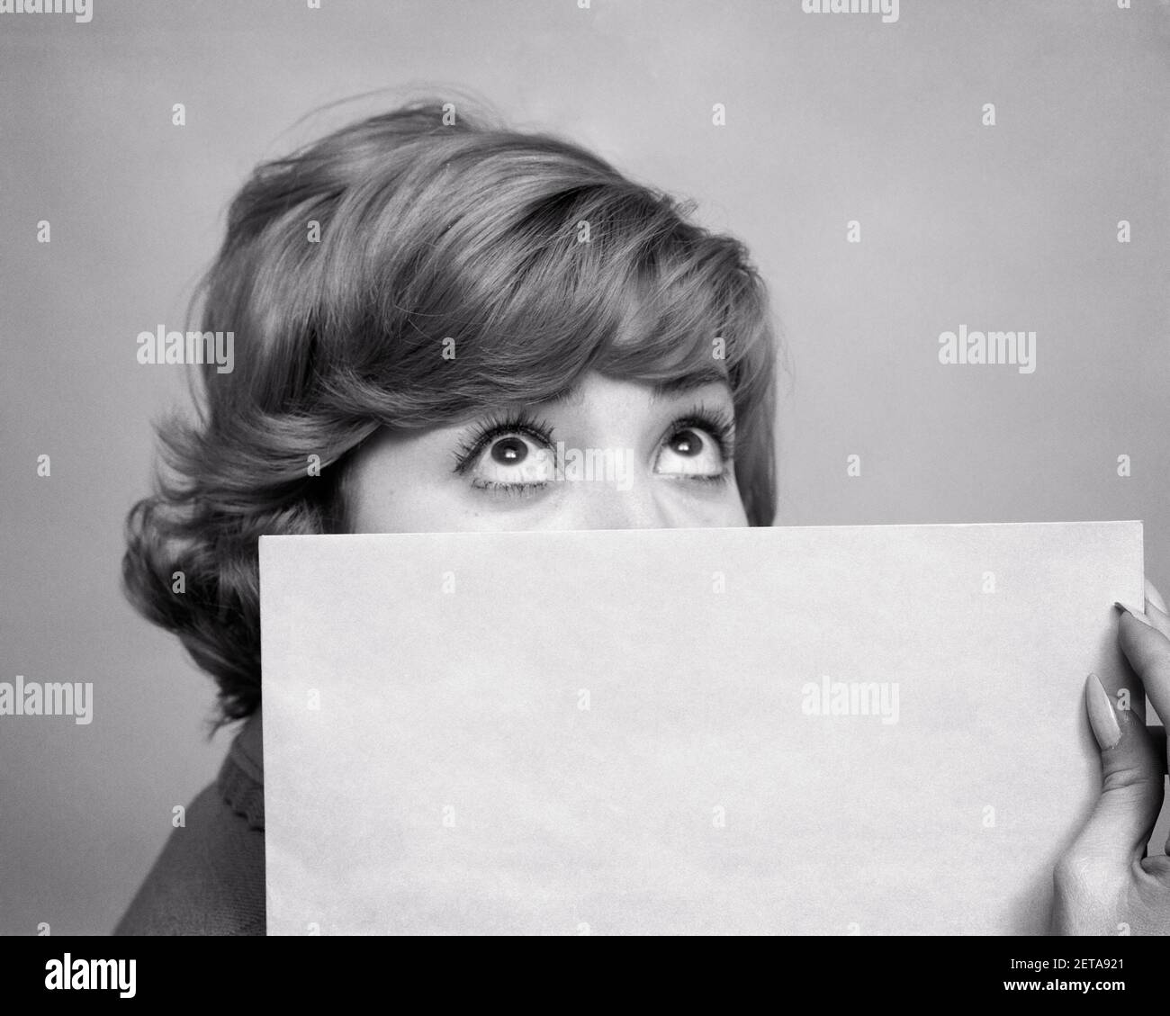1960s WOMAN GREAT BIG EYES LOOKING UP OVER THE TOP OF A BLANK SIGN CARD HIDING THE LOWER PART OF HER FACE - g3702 DUN001 HARS COPY SPACE LADIES LOWER PERSONS SYMBOLS B&W BUG-EYED HUMOROUS HEAD AND SHOULDERS ADVERTISEMENT AD COMICAL PART OPPORTUNITY OCCUPATIONS CONCEPT CONNECTION CONCEPTUAL COMEDY SYMBOLIC WIDE-EYED CONCEPTS IDEAS LOOKING UP UNMARKED YOUNG ADULT WOMAN BLACK AND WHITE CAUCASIAN ETHNICITY MESSAGE OLD FASHIONED REPRESENTATION UNUSED Stock Photo