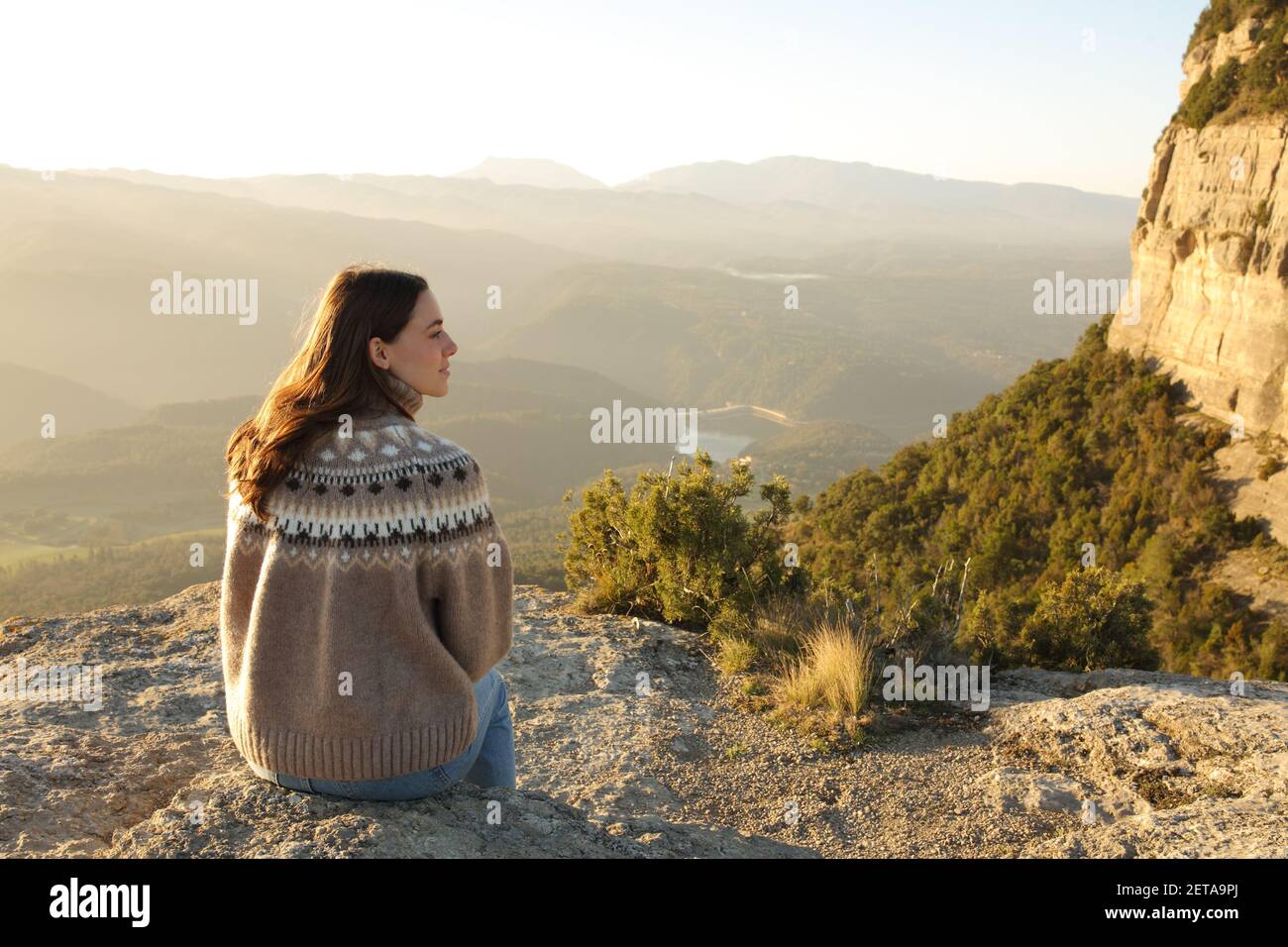 Back view portrait of a woman sitting in a cliff contemplating views at sunset Stock Photo