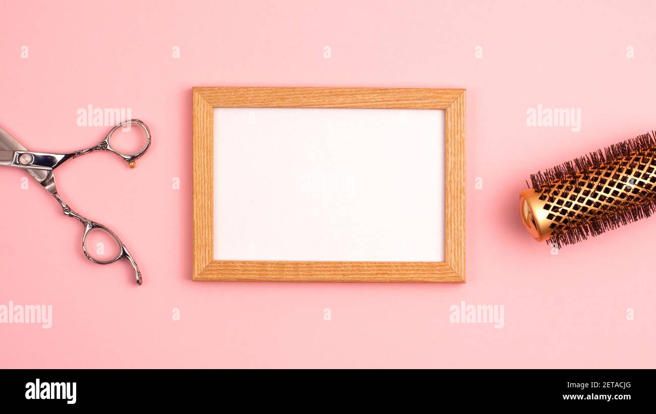 Wooden picture frame mockup flat lay of professional hair cutting shears, gold round hair brush for styling template on pink background with copy spac Stock Photo