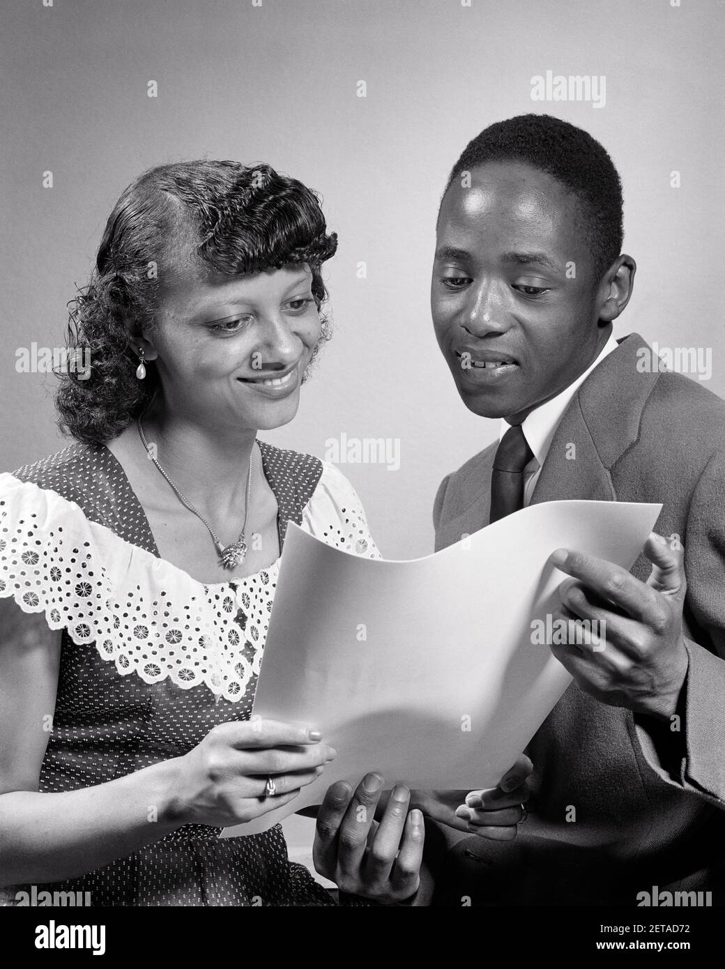 1950s AFRICAN-AMERICAN COUPLE READING DOCUMENT CERTIFICATE DIPLOMA WOMAN  WEARING DOTTED SWISS DRESS BIG COLLAR MAN SUIT AND TIE - n696 HAR001 HARS  STYLE COMMUNICATION YOUNG ADULT INFORMATION LIFESTYLE SATISFACTION FEMALES  PROUD DIPLOMA