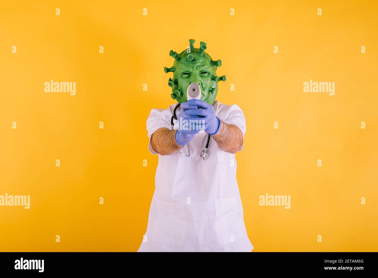 Doctor - nurse in coronavirus disguise wearing latex mask - covid-19 virus pointing with an electronic thermometer on yellow background. Coronavirus c Stock Photo