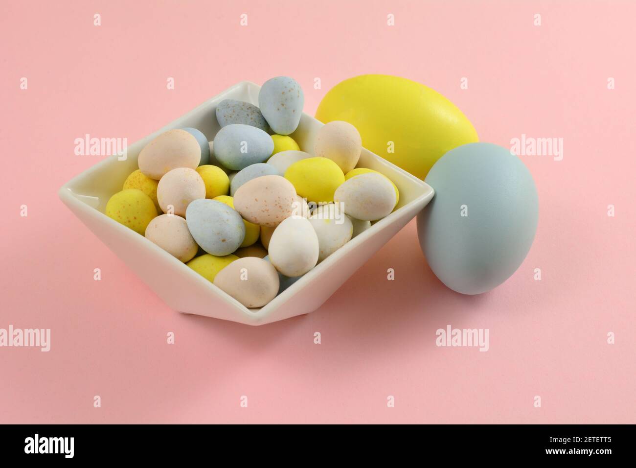 Pastel colored candy Easter eggs in white dessert bowl with two painted Easter eggs on pink background Stock Photo