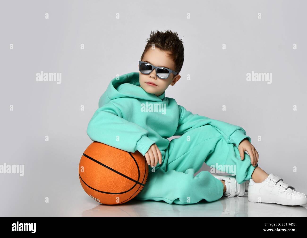 Cool kid boy in modern green, mint color sportswear hoodie, pants and white sneakers sits with basketball looking back Stock Photo