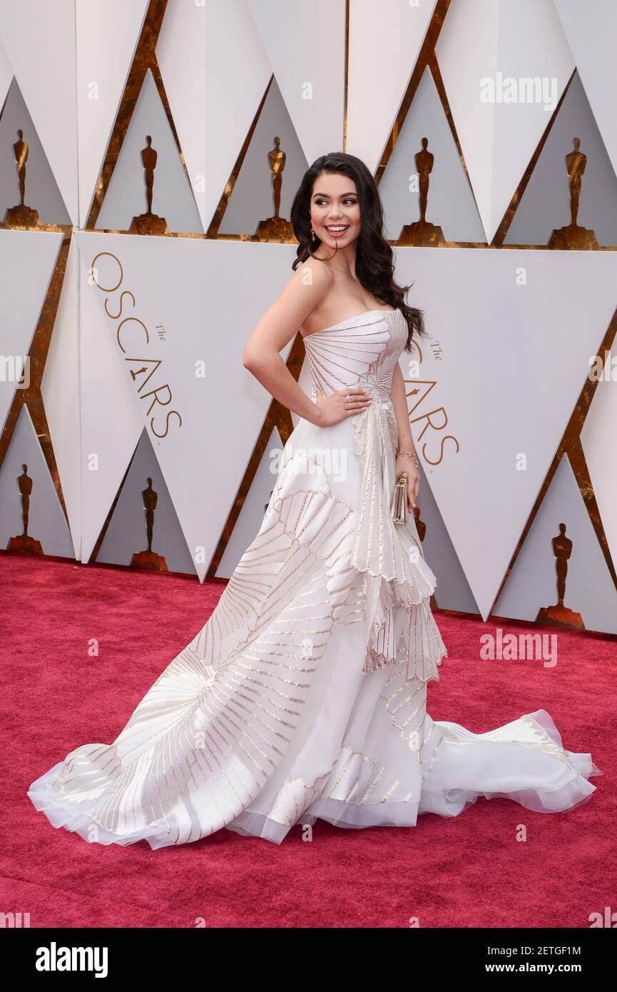 Auli'i Cravalho wearing a red Valentino lace dress with an Irene Neuwirth  crown and earrings and an ACLU blue ribbon walking the red carpet during  the 89th Academy Awards ceremony, presented by