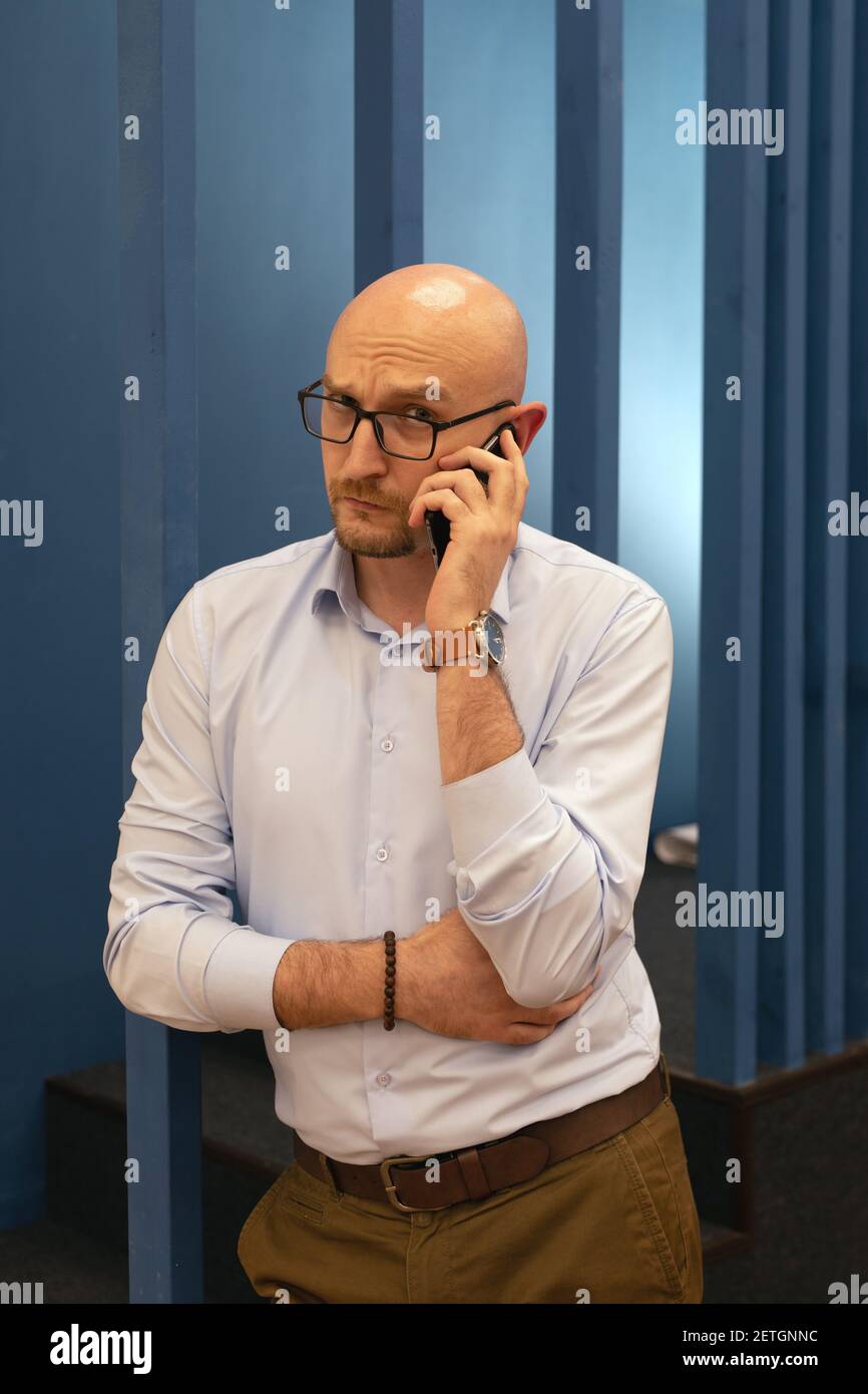 Adult bald man with glasses looks at camera, calls on mobile phone. Serious businessman solves problems Stock Photo