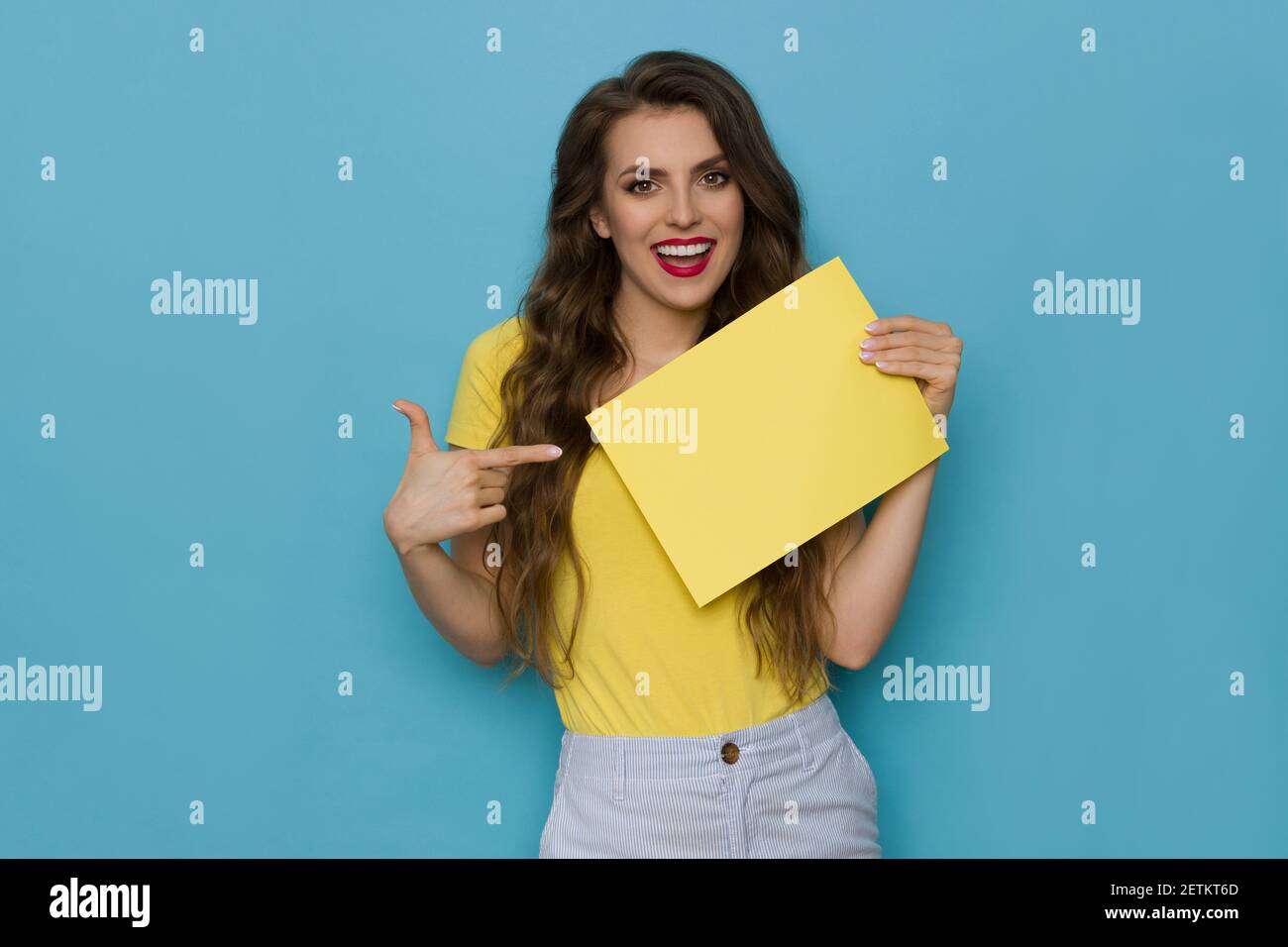 Happy casual young woman is holding empty yellow sheet of paper, pointing at it and talking. Front view. Waist up studio shot on blue background. Stock Photo