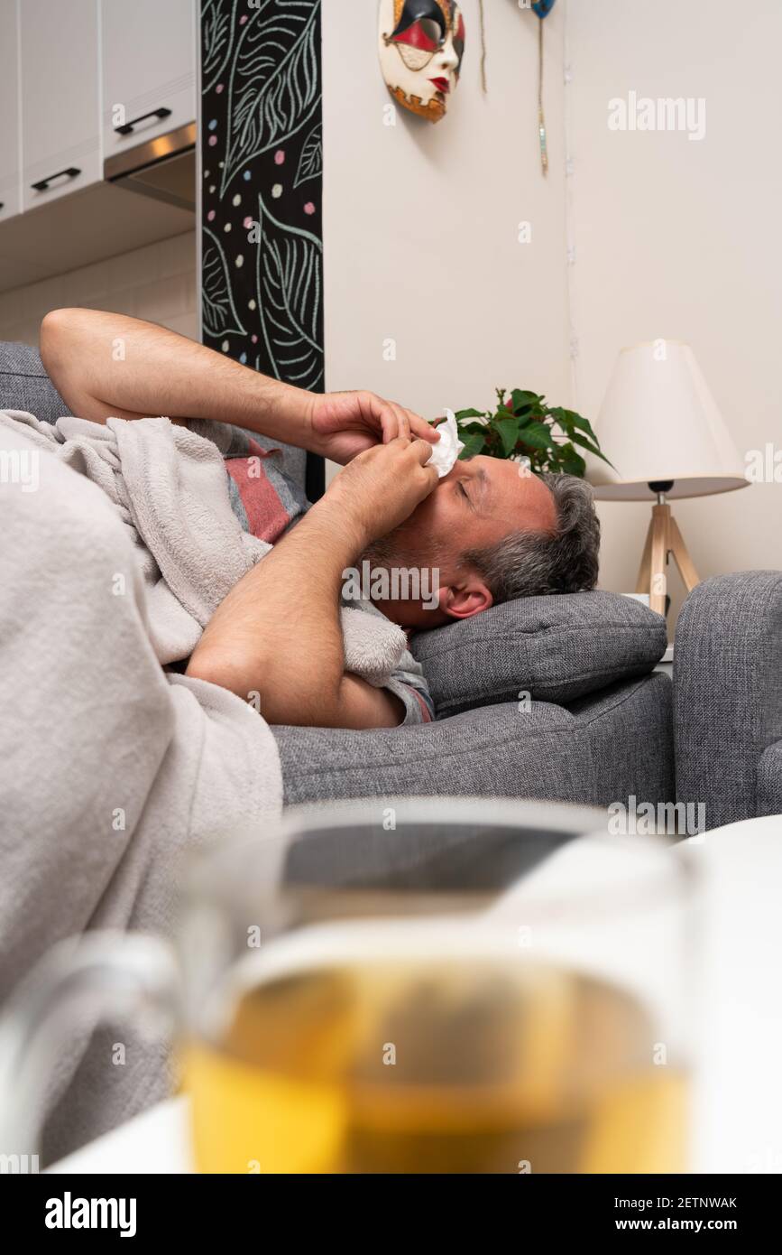 Sick adult man model with sinusitis cold flu symptoms blowing nose using tissue resting on sofa at home with cup of tea on table as pandemic concept Stock Photo