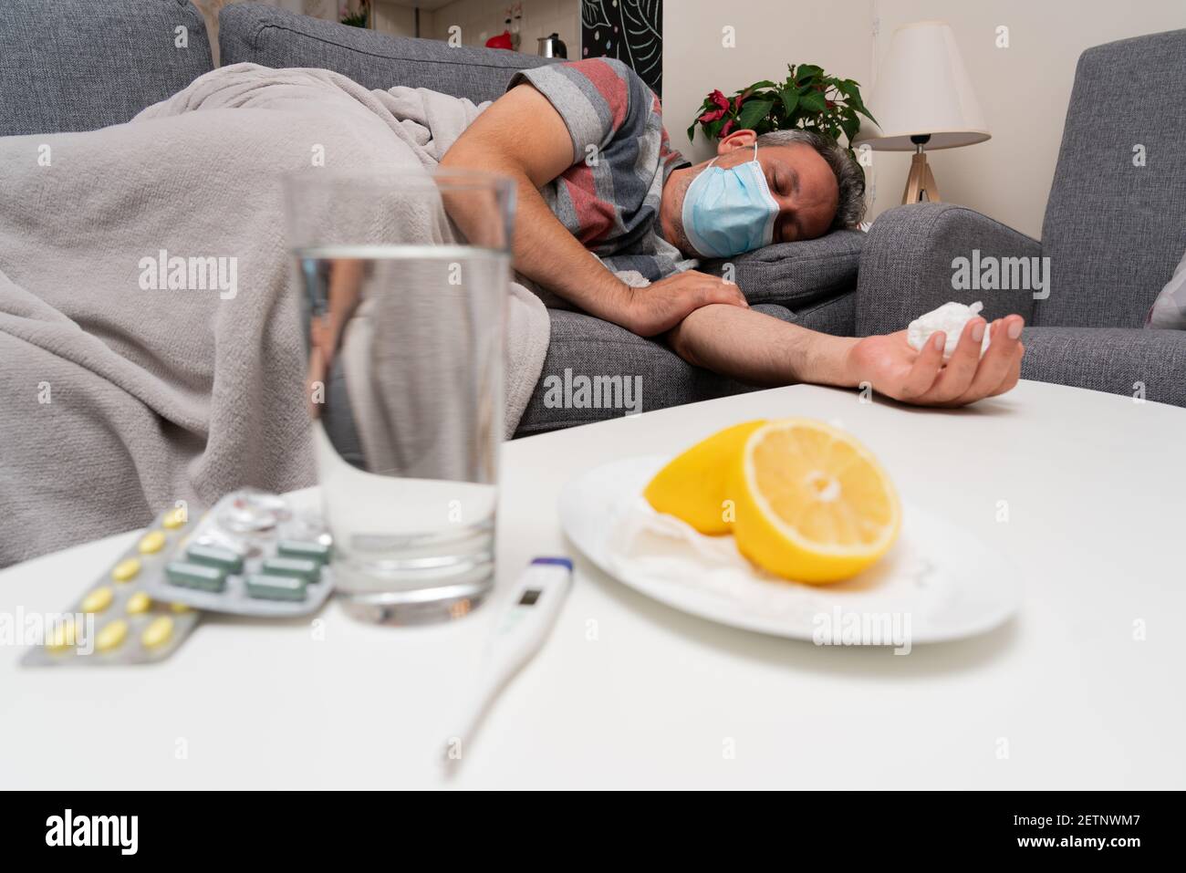 Sick male model wearing surgical or medical protective mask to prevent contamination sleeping on sofa cold sars covid19 treatment pills water thermome Stock Photo