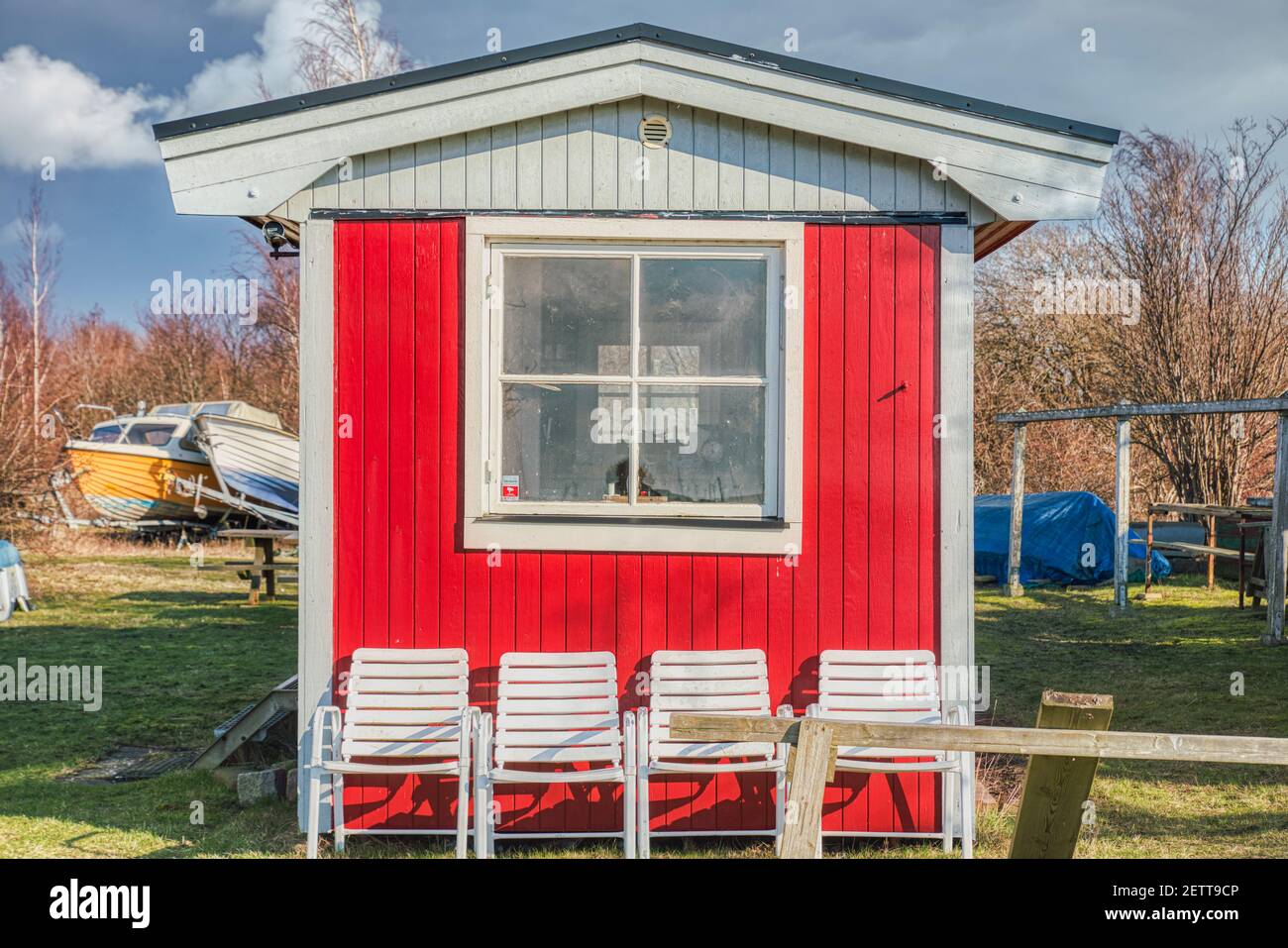 A typical Swedish red summerhouse with a square window and white chairs. The bare exterior has a simple aspect with its wooden facade and slanted roof Stock Photo
