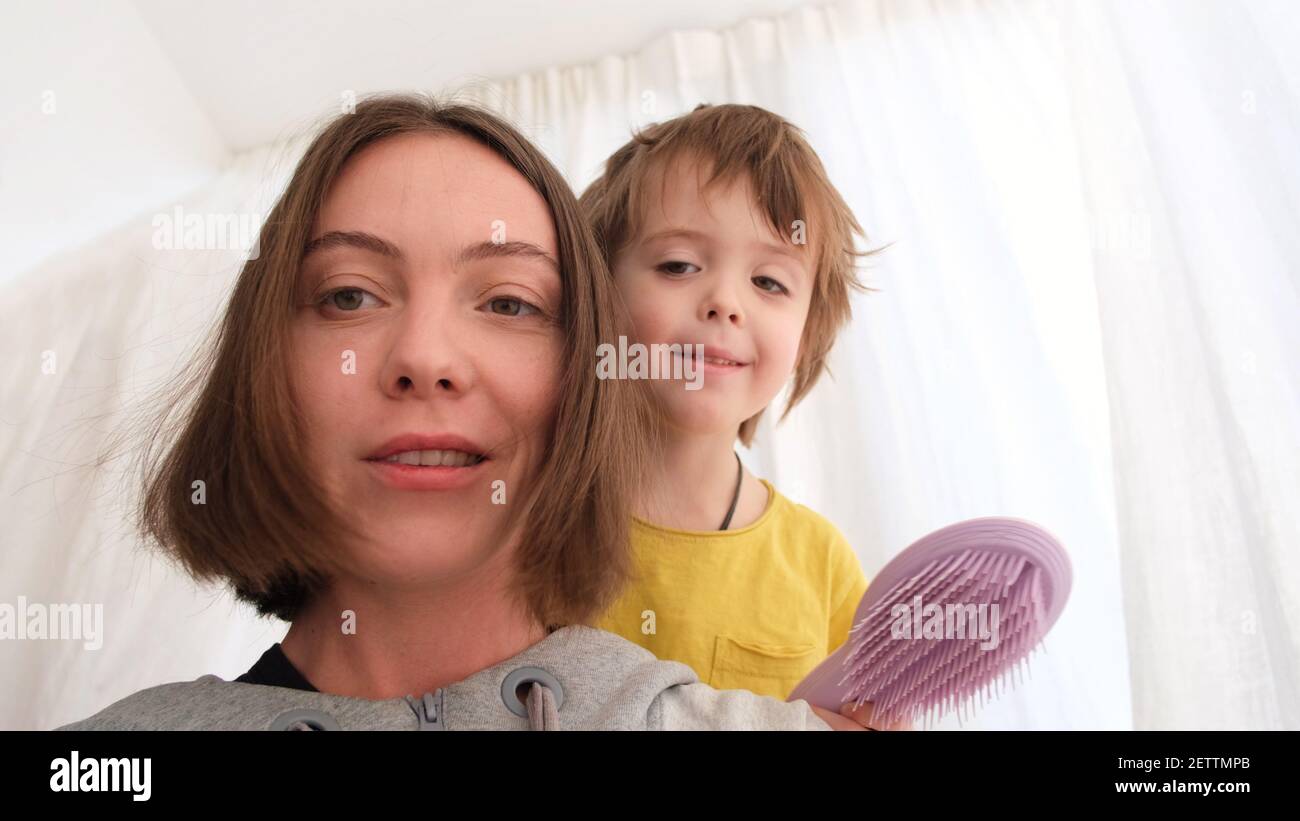 Cute little boy brushes happy mother short hair looking at each other during videocall near window in light room closeup Stock Photo