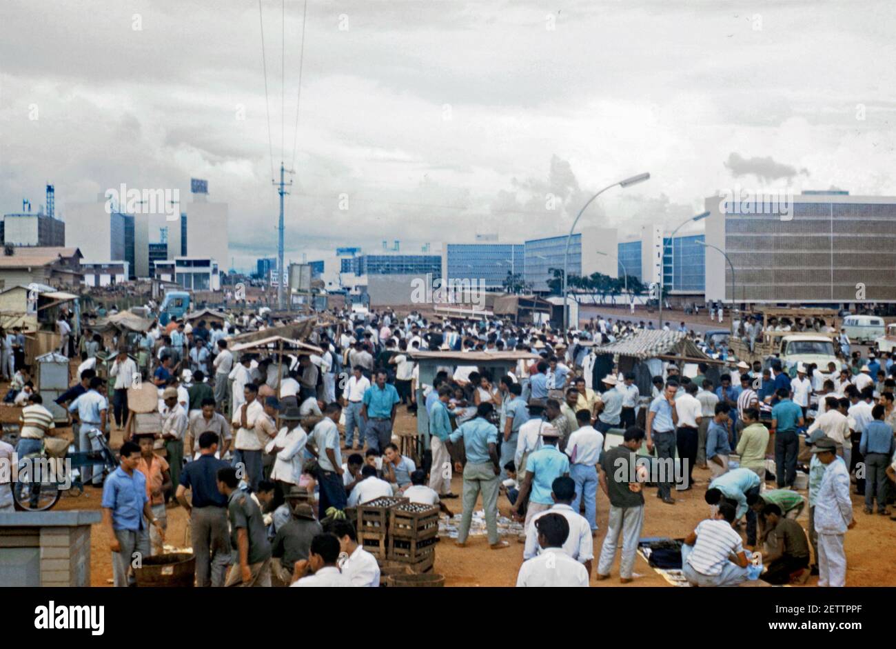 Old meets new – a traditional, busy Sunday street market in the modern setting of Brasília, Brazil 1961. The dull conditions haven’t put off the crowds, very much male dominated. The architecture of the unfinished modern city is seen in the background. Brasília is the federal capital of Brazil. The city is located in the Brazilian highlands in the country's centre-west region. Brasília was a planned city developed by Lúcio Costa, Oscar Niemeyer and Joaquim Cardozo in 1956. This image is from an old amateur 35mm colour transparency. Stock Photo