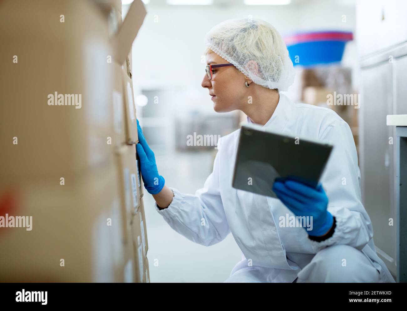 Close up view of a young female beautiful focused worker in sterile cloths inspecting a stack of boxes while holding the tablet. Stock Photo