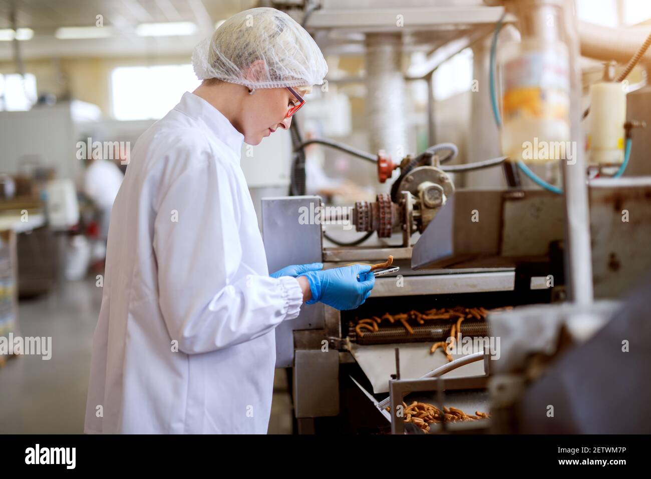 Close up view of a young female worried worker in sterile cloths inspecting salt sticks taken from the food snack production line. Stock Photo
