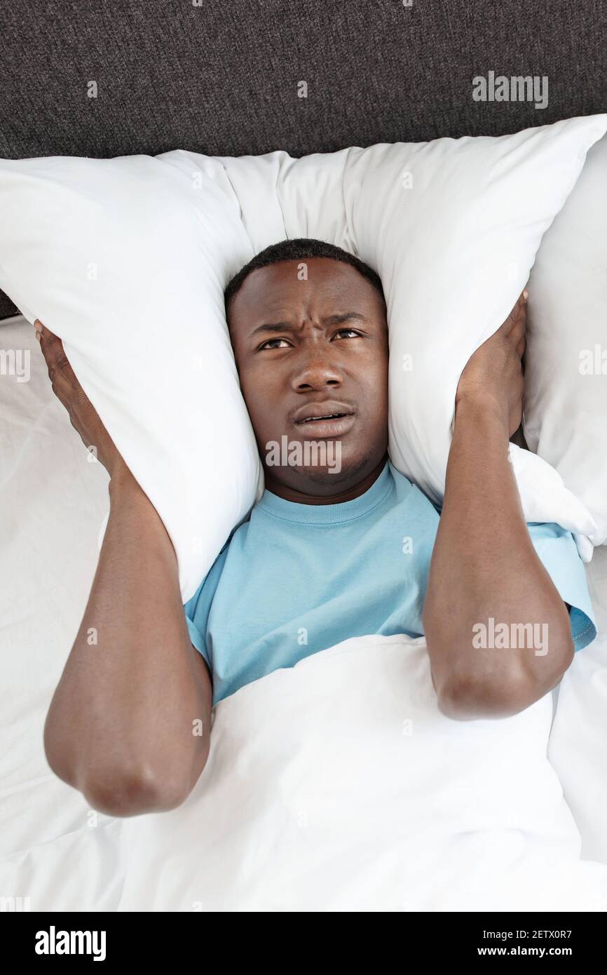 Upset tired young african american man freaking out, covers his ears with pillow so as not to hear noise in bedroom Stock Photo