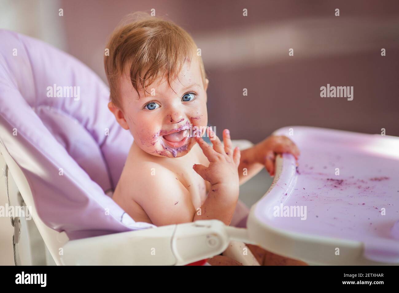 Adorable baby eating on baby chair and making a mess. Little baby girl eating food. Cute baby with a funny expression on his face. Stock Photo