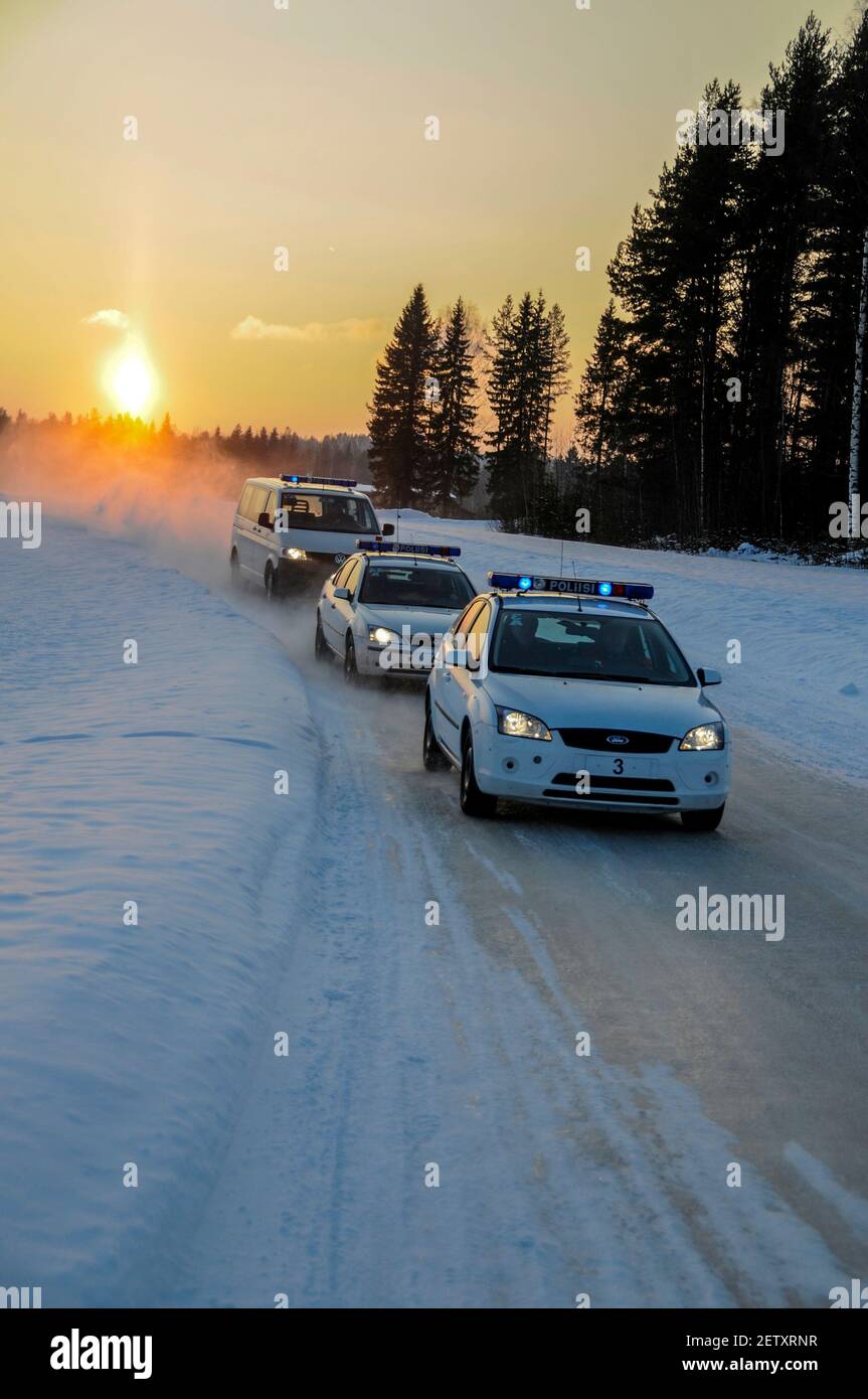 A convoy of blue flashing Finnish Traffic Police car speeds along a snow-covered road as the winter sunsets at Pieksamaki in Finland.  The Police were Stock Photo