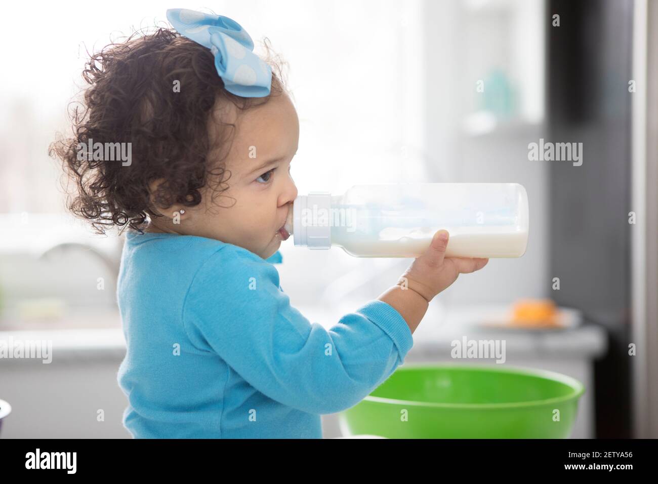 A little toddler girl drinks milk from a plastic bottle in a bright