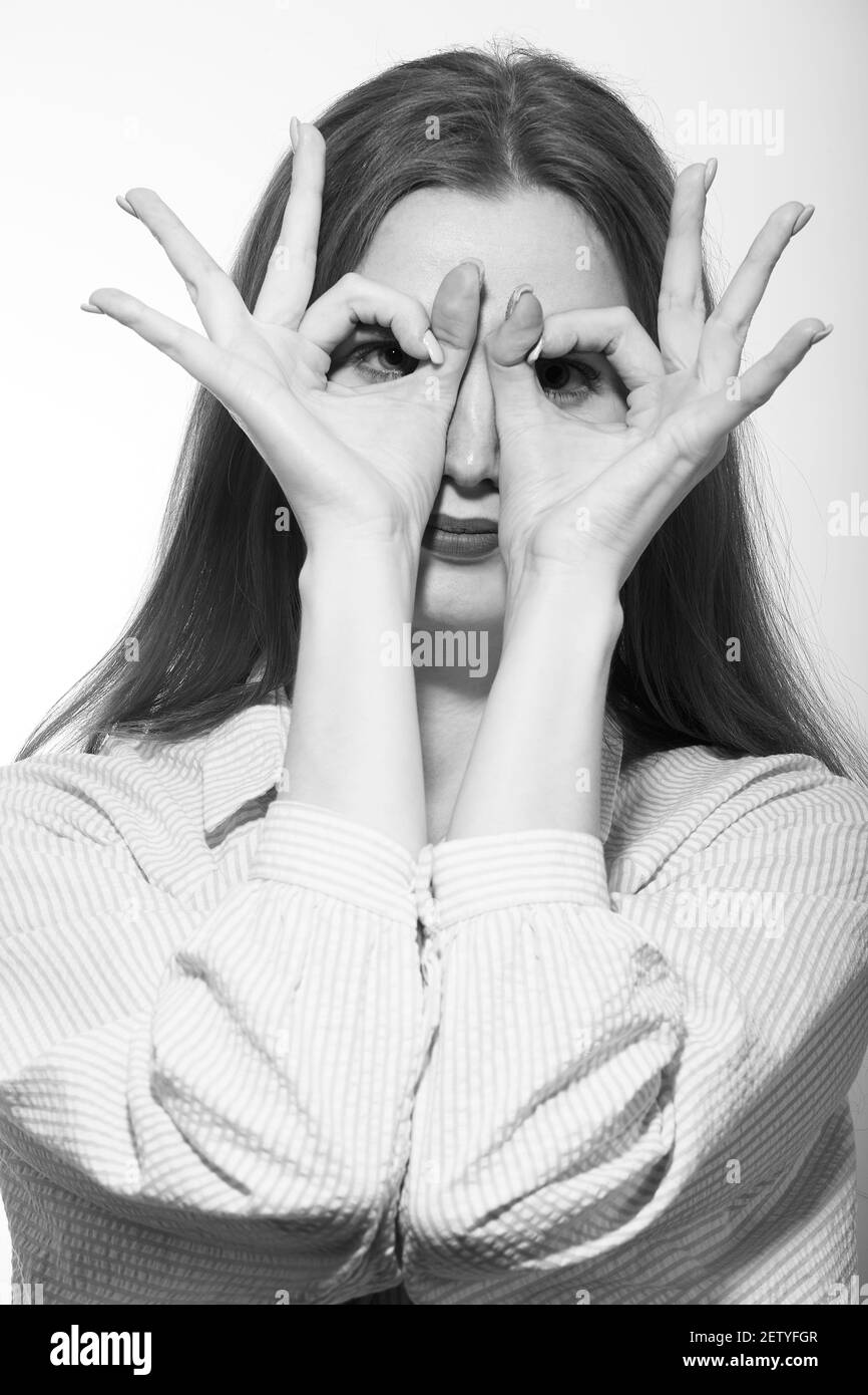 serious young woman looking at camera through her fingers with eyeglasses gesture on white background, monochrome Stock Photo
