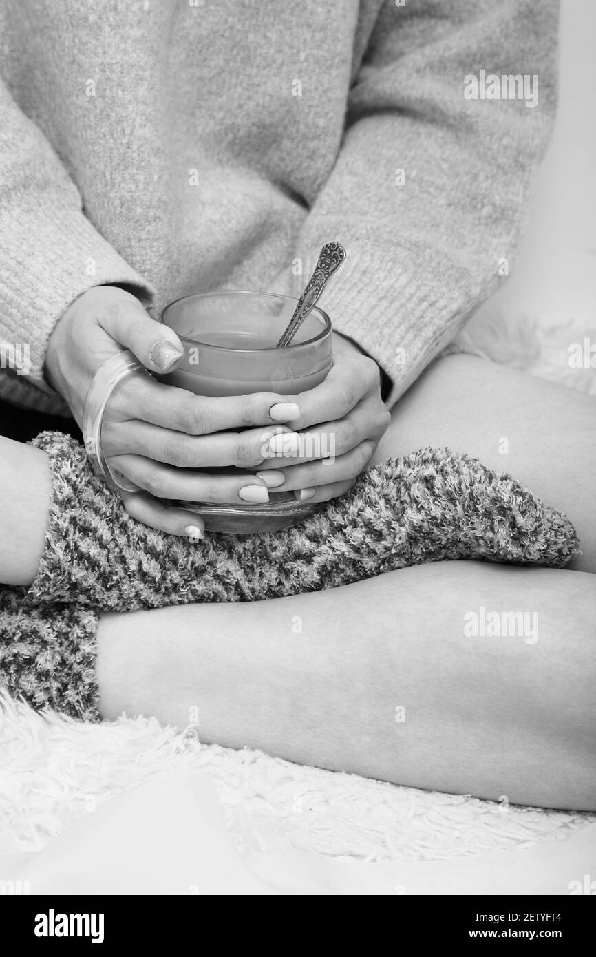 unrecognizable young woman in sweater and woolen socks with cup of hot tea, closeup hands and feet, monochrome Stock Photo