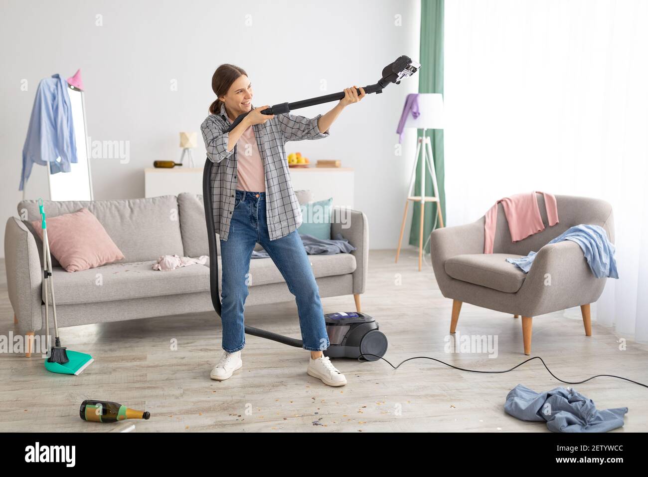 Positive young lady holding vacuum cleaner like rifle, ready to perform cleanup at messy room after party, copy space Stock Photo