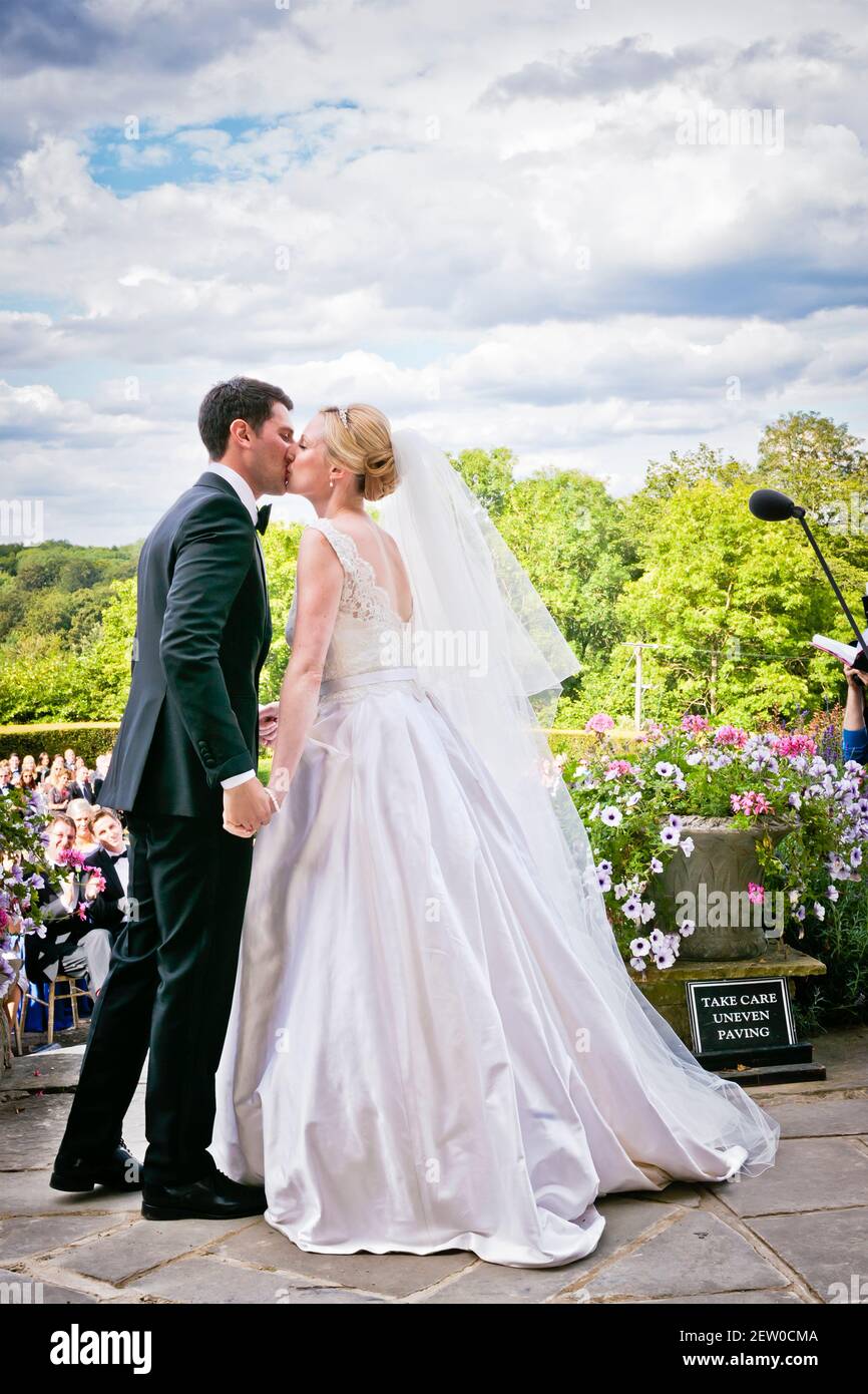 Bride and Bridegroom kissing. They are overlooked by their guests who are all seated withing a lower area of the garden. Stock Photo