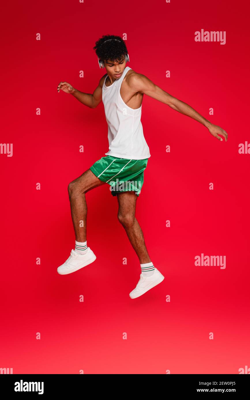 full length of curly african american man in wireless headphones jumping on red Stock Photo