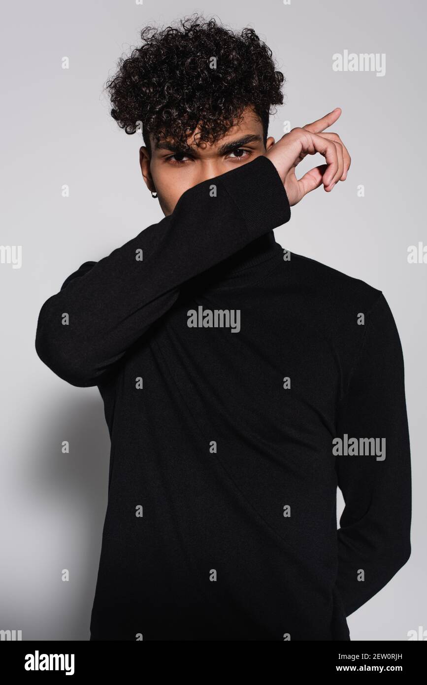 curly african american man covering face with hand while posing on grey ...