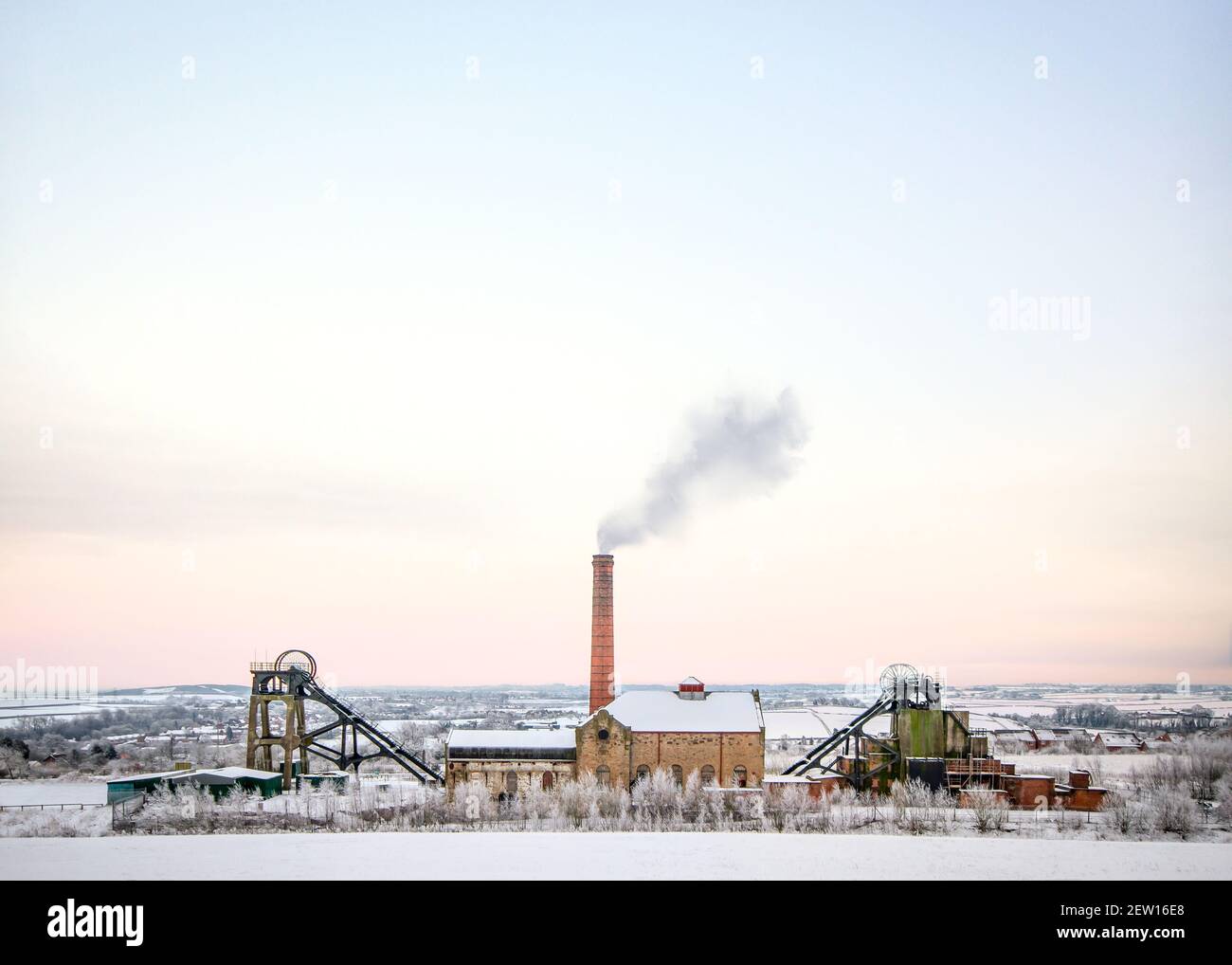 Old working coal board mine in countryside landscape with smoke from chimney of colliery engine house winding wheel. Under snowfall Christmas winter Stock Photo