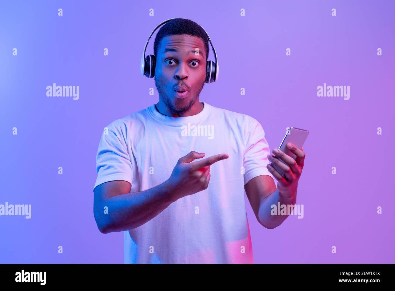 Cool App. Surprised Black Guy In Wireless Headphones Pointing At Smartphone Stock Photo