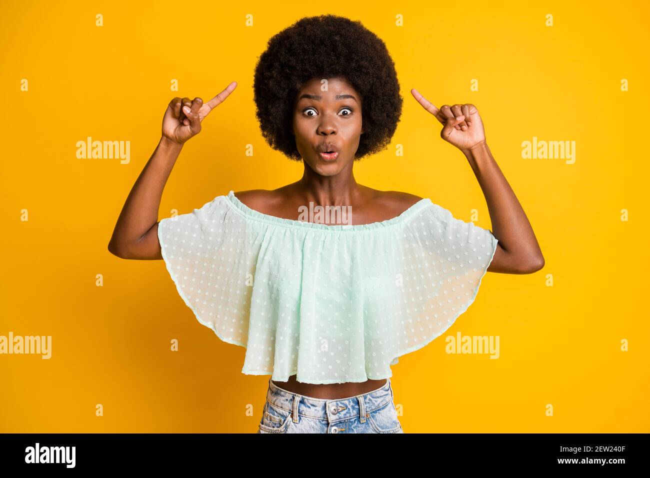 Photo portrait of shocked woman pointing two fingers at afro wig isolated on vivid yellow colored background Stock Photo