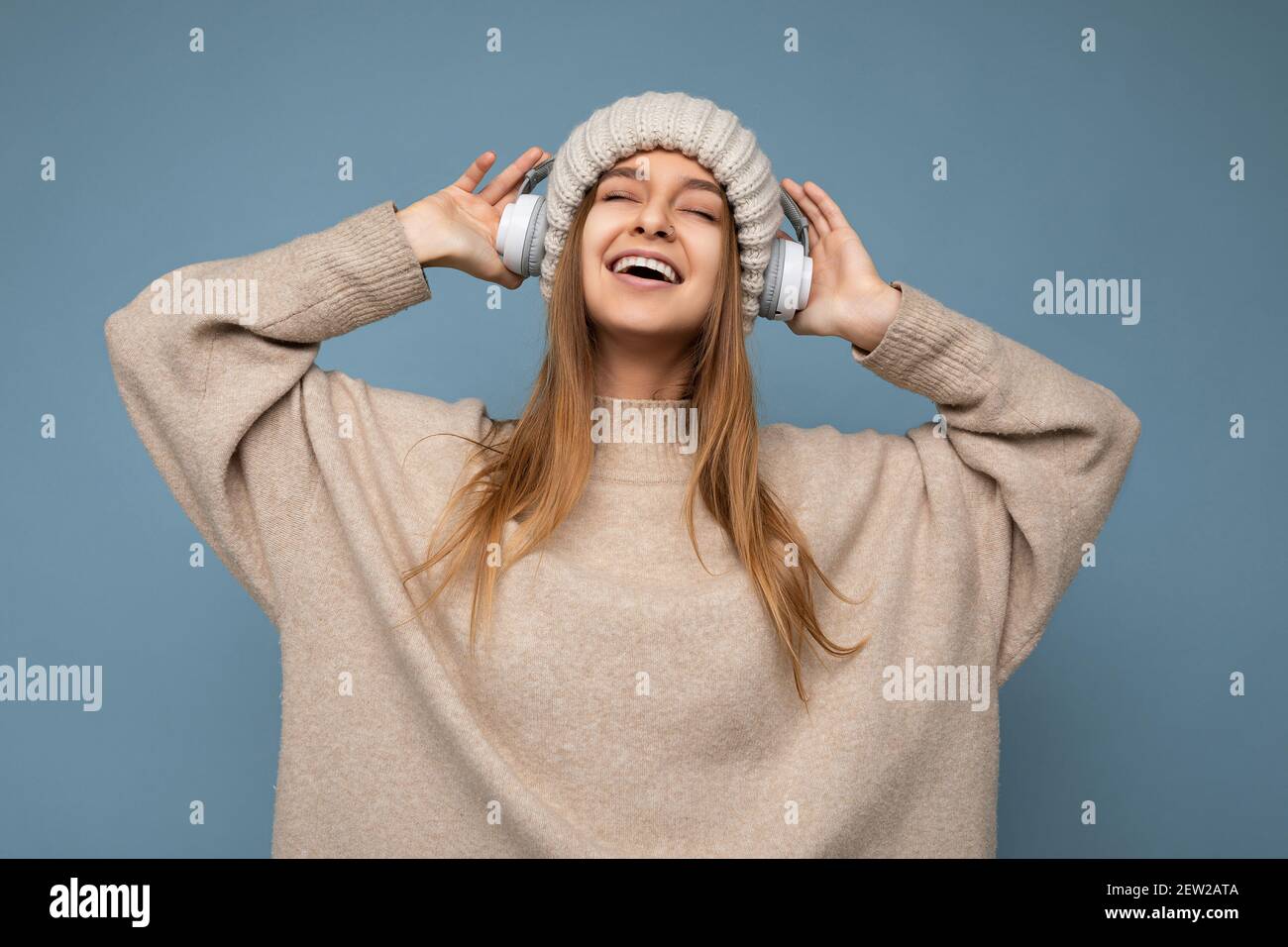 Photo of beautiful happy smiling young blonde woman wearing beige winter sweater and hat isolated over blue background wearing white wireless Stock Photo
