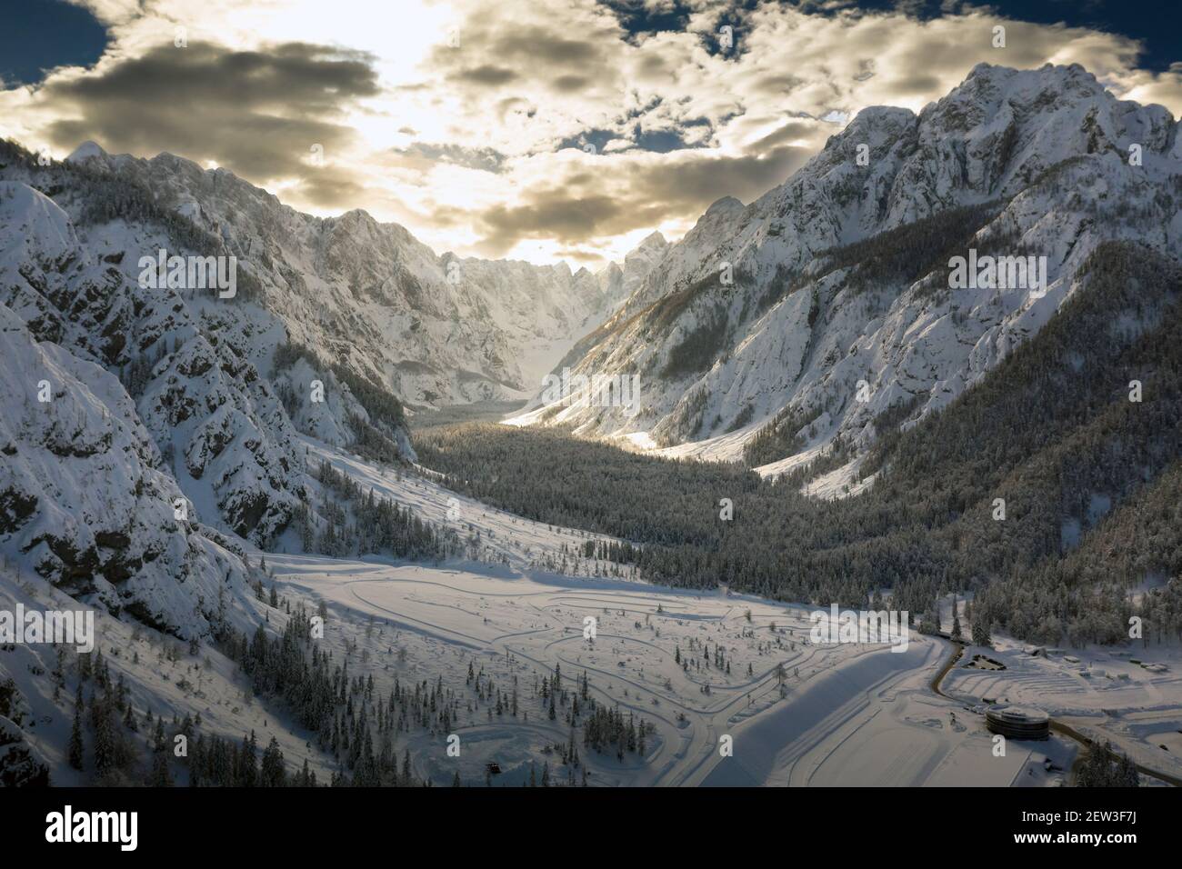 Aerial flight over the mountain valley in winter covered in snow. Cinematic drone view with sunlight breaking through clouds. Stock Photo