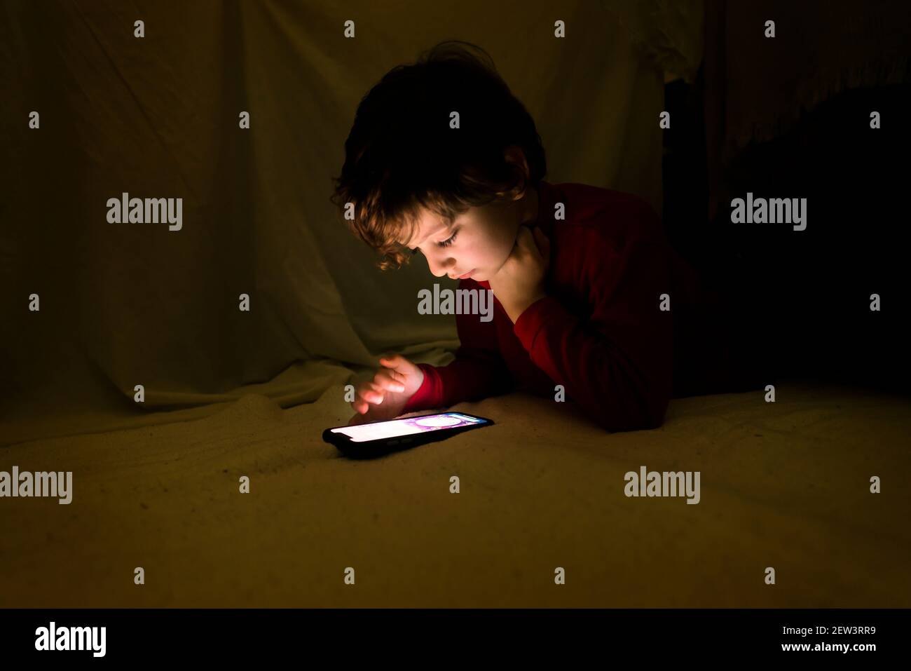 Child watching a smartphone, lying on a blanket, in a makeshift tent in his living room. Home lifestyle concept. Stock Photo