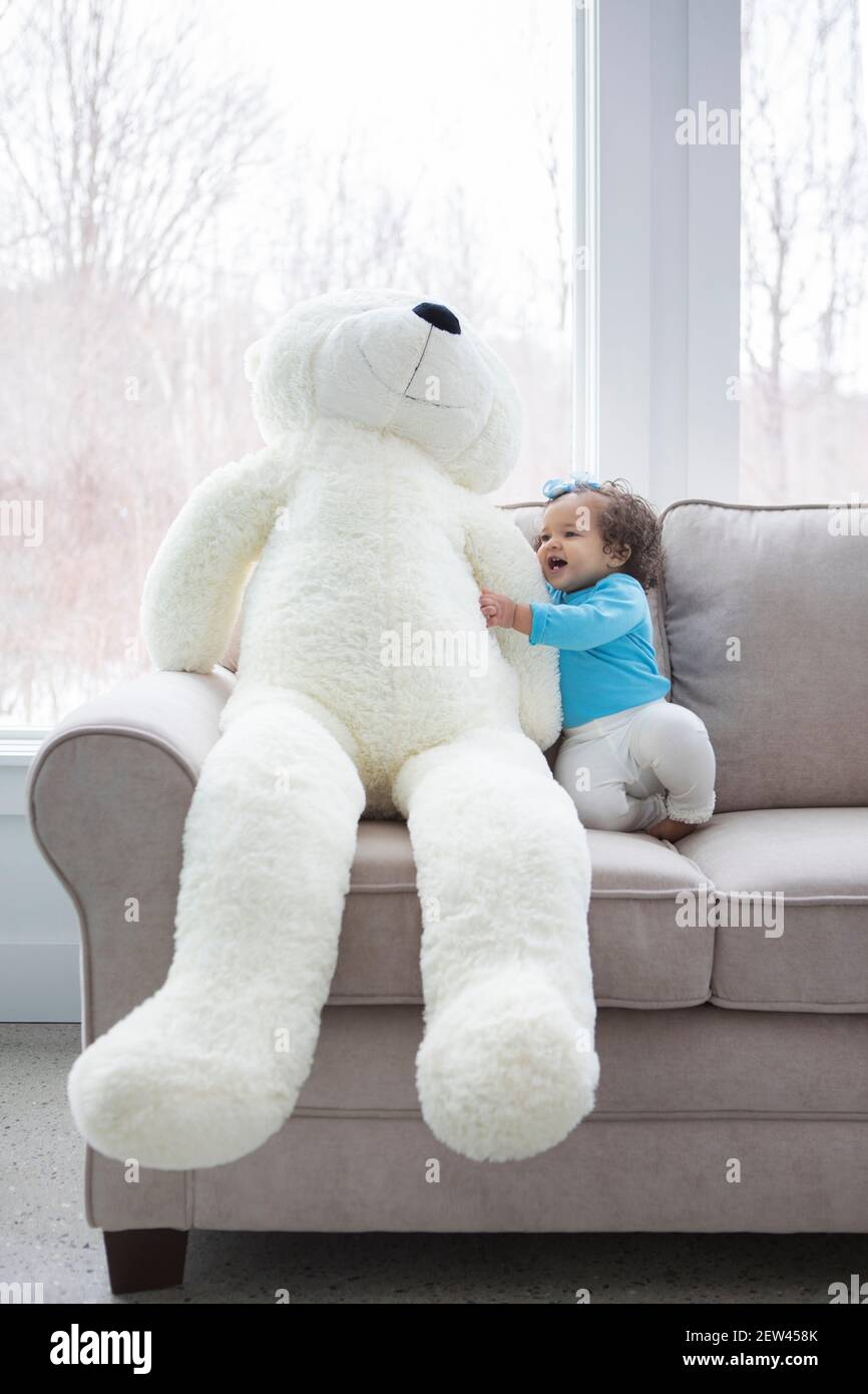 A little girl sits on a sofa with a giant white teddy bear. Stock Photo