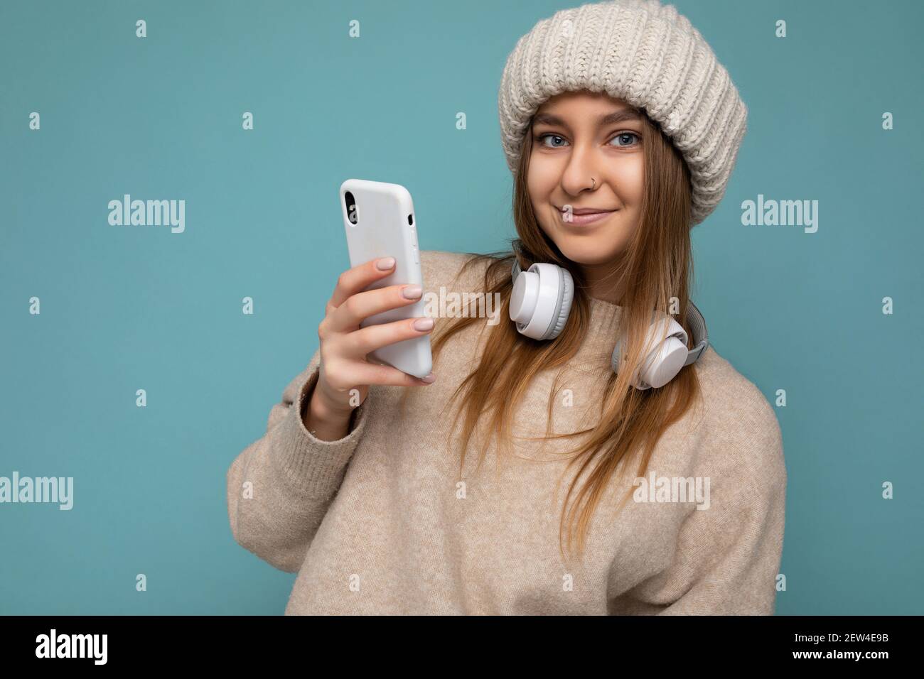 Closeup Photo of beautiful happy smiling young dark blonde woman wearing beige stylish sweater and knitted beige winter hat isolated on blue Stock Photo