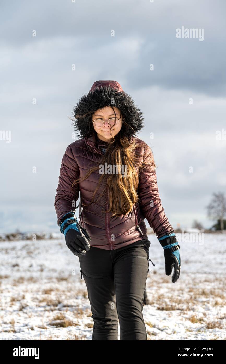 A teenage girl in a winter jacket with a hood in a cold winter landscape. Snow and a cloudy sky in the background Stock Photo