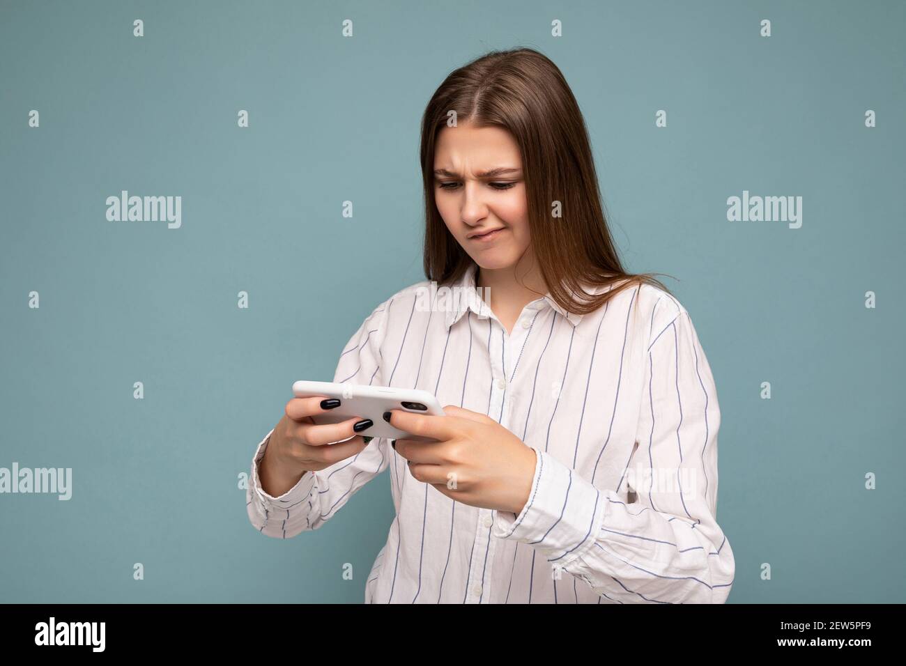 Concentrated attractive young blonde woman wearing casual white shirt isolated over blue background wall holding smartphone and playing online games Stock Photo