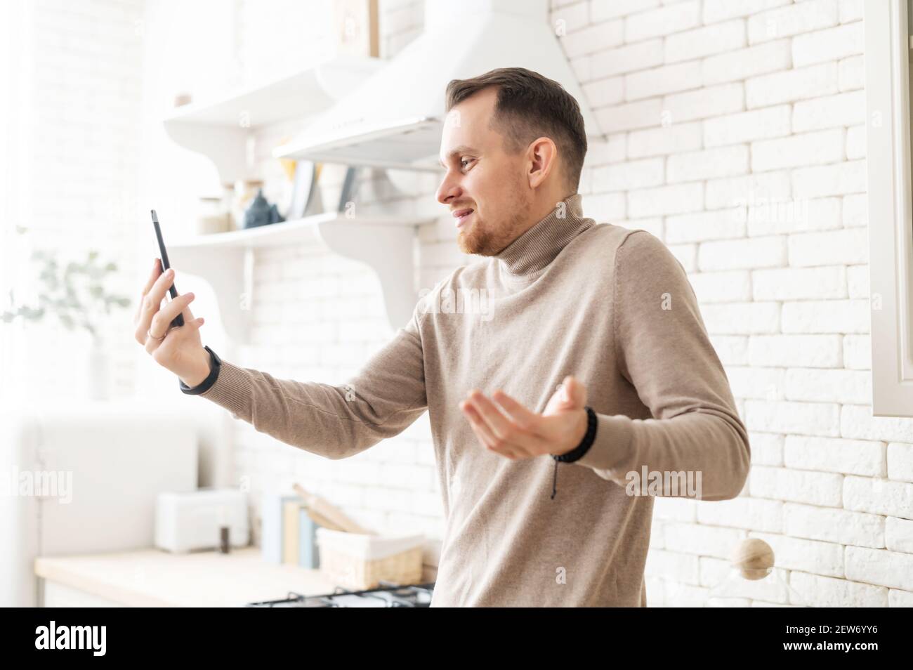 Young masculine man using smartphone for video call to talk to his friends  remotely, shrugs, making online call to his wife or business partner,  speaking with family on internet, using high speed