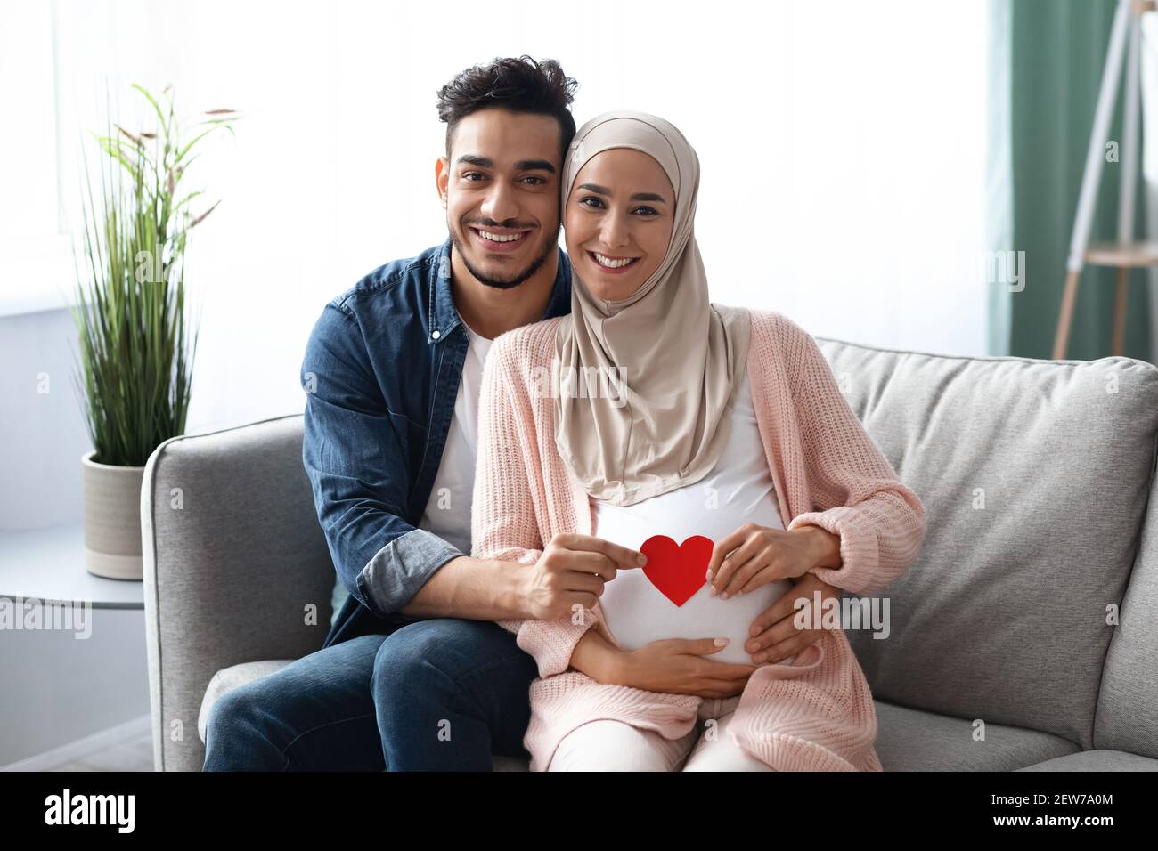 Happy Expectation. Muslim Couple Holding Red Paper Heart Near Woman's Pregnant Belly Stock Photo