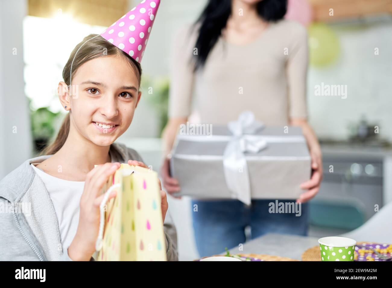 Pretty teenaged latin girl smiling at camera while holding gift bag, receiving presents, celebrating birthday with mother at home. Celebration, childhood concept. Selective focus Stock Photo