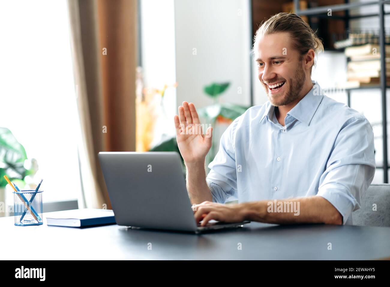 Attractive freelancer guy using laptop, have a video call, communicate with colleagues or friends by video conference, waving hand and smiling. Handsome caucasian man sits at the desk, working from Stock Photo