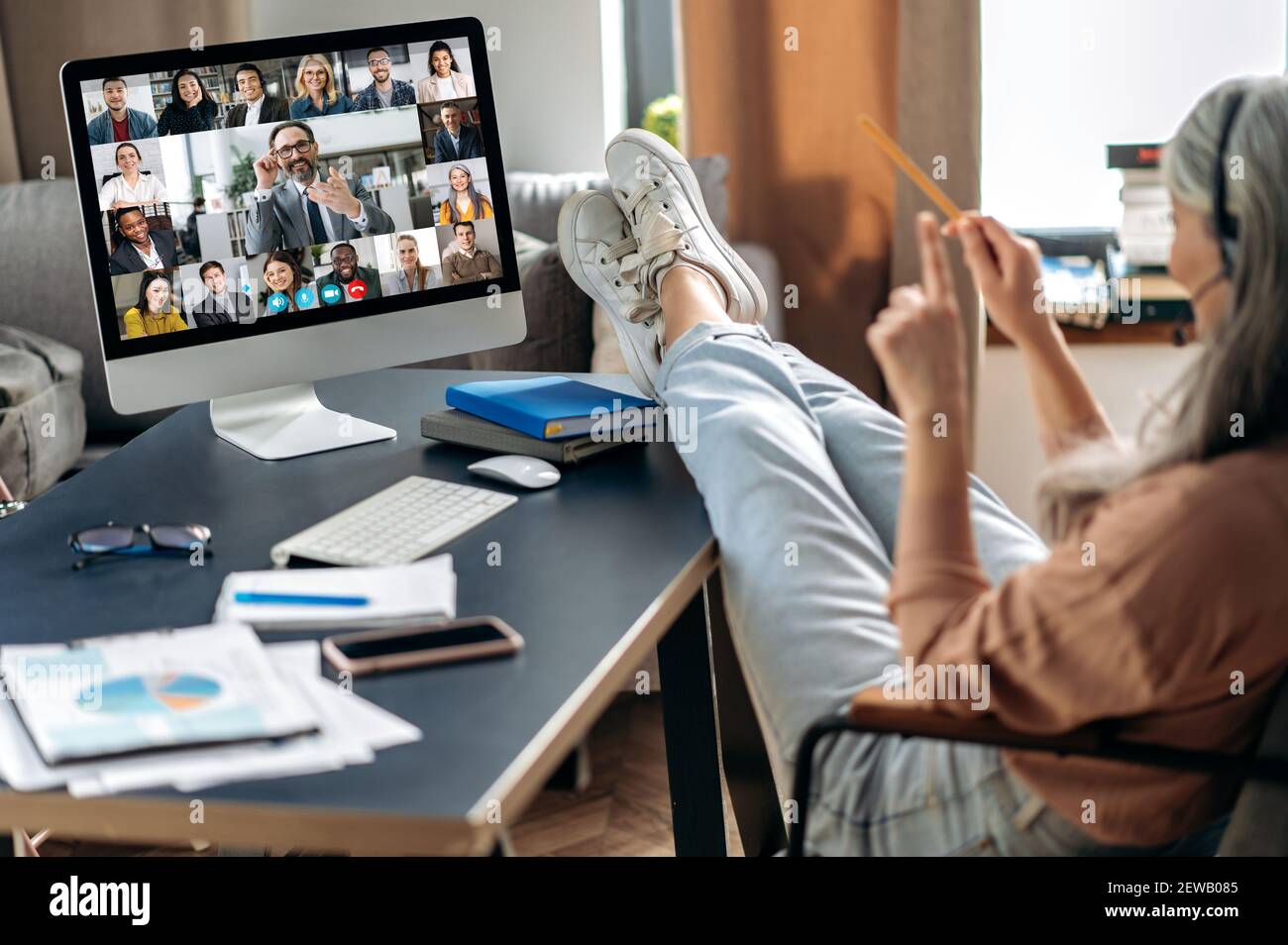 Remote work from home. Modern aged gray-haired woman communicate with coworkers online via video call uses app and computer, sitting at her home office, on the screen many different people Stock Photo